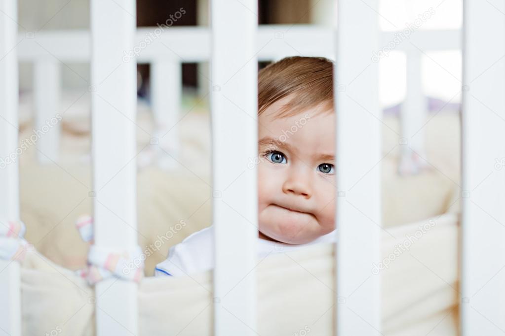 Little cute adorable little blond boy in a striped bodykit sitting with pursed lips behind bars white cot