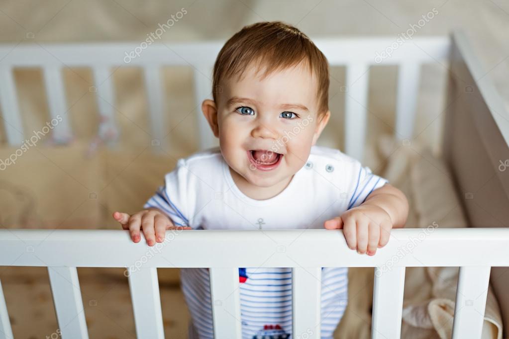 Little cute adorable little blond boy in a striped bodykit is in the nursery with white crib and laughs
