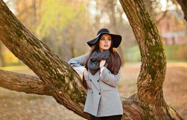 Bir gri ceket ve siyah şapka parkta sonbahar manzara arka plan üzerinde bir bankta oturmuş, parlak düz saç ile çok güzel bir genç esmer kadın portresi