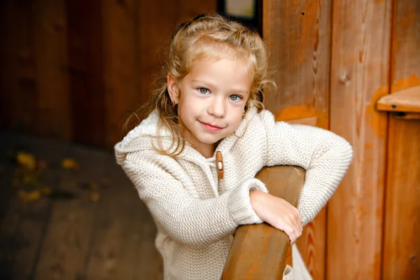 Adorable little curly blond girl in beige knitted sweater smiles  slyly, against the background of a wooden house in autumn — Stock Photo, Image