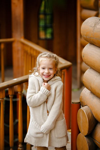 Schattig klein krullend blond meisje in beige gebreide trui glimlacht sluw, tegen de achtergrond van een houten huis in de herfst — Stockfoto
