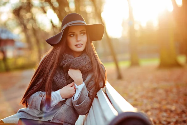 Портрет очень красивой молодой брюнетки с блестящими прямыми волосами в сером пальто и черной шляпе, сидящей на скамейке на фоне осеннего пейзажа в парке — стоковое фото