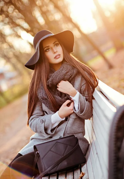 Ritratto di una giovane donna bruna molto bella con capelli lisci lucidi in cappotto grigio e cappello nero, seduta su una panchina sullo sfondo del paesaggio autunnale nel parco — Foto Stock