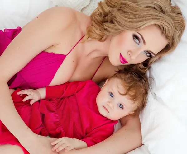 Стройная блондинка-мама в красном платье лежит на кровати рядом с дочкой — стоковое фото