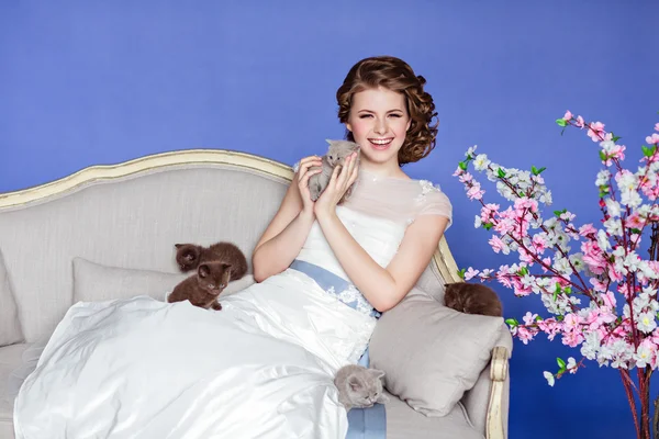 Gülümseyen ve onun beş küçük kedi yavrusu tutan sevimli ve güzel kız beyaz elbise sakura çiçekli mavi bir arka plan kanepede oturan — Stok fotoğraf