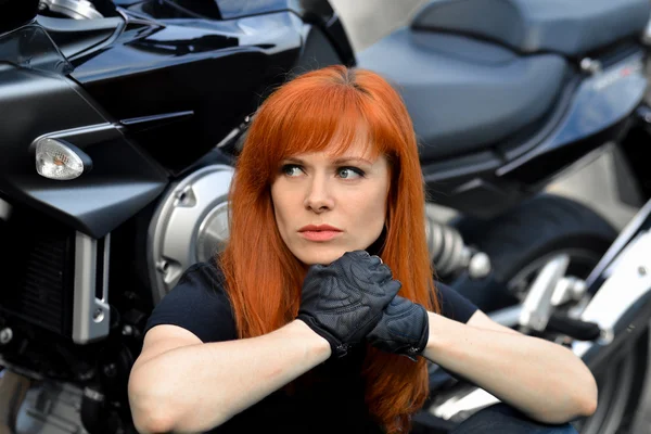 Deri eldivenve serin, modern bisiklet, motosiklet ile tehlikeli bir görünüm ile meraklı, ciddi, kızıl saçlı kız. Profesyonel motorcu. Ciddi, güçlü, bisikletçilerin düşünen lideri bisikletin yanında oturup kenara bakın