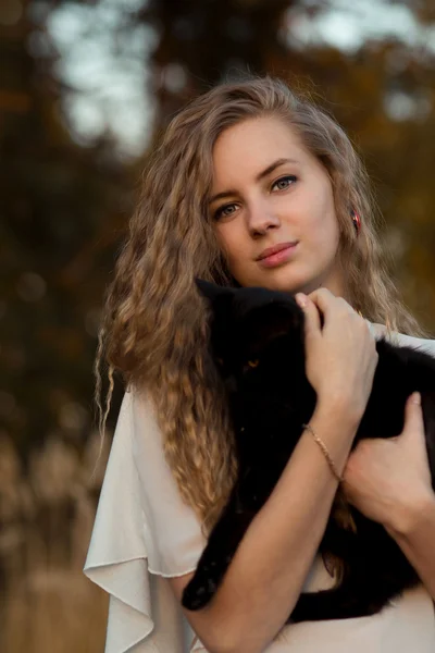 Nice girl with her favourite pet - black cat.Beautiful happy smiling blonde girl hold black cat in hands.Cute girl with long curly hair hug little, small black cat.Cute, friendly, kind girl love animals, pets, cats and dogs.Cat is friend to people.