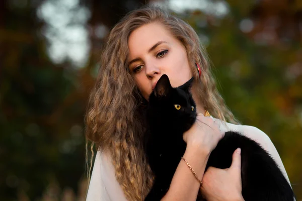Nice girl with her pet  black cat.Beautiful happy healthy smile.Beautiful happy smiling blonde girl hold black cat in hands.Cheerful,happy and smiling girl with perfect smile caress and held black cat in hands in summer,spring,autumn forest,outdoors.