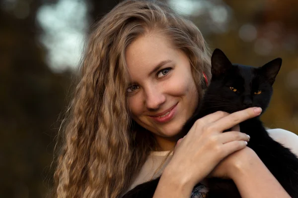 Nice girl with her favourite pet - black cat.Beautiful happy smiling blonde girl hold black cat in hands.Cute girl with long curly hair hug little, small black cat.Cute, friendly, kind girl love animals, pets, cats and dogs.Cat is friend to people. — Stock fotografie