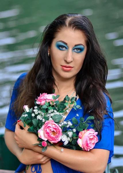 Vacker flicka med kreativ blå makeup och bukett blommor står nära den blå floden, sjö, vatten. — Stockfoto