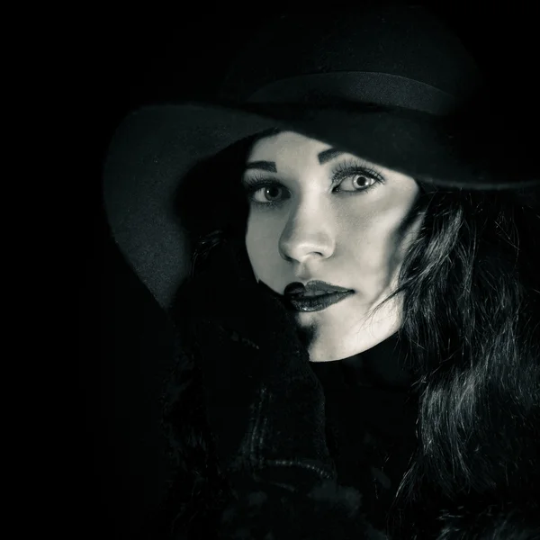 Closeup fashionable portrait of beautiful, pretty girl in black hat. Attractive woman with bright amazing green eyes, red lips looking at camera. Black and white studio portrait of an classic, vintage girl in black hat on black bakcground.