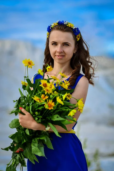 Gyönyörű aranyos lány, hosszú, kék ruhában, a csokor sárga virágok koszorú a virágok. Portré gyönyörű, vonzó, aranyos, kedves, csodálatos ukrán lány, gyönyörű csokor virág: sárga százszorszépek. Vidám lány, mosolygó lány. — Stock Fotó