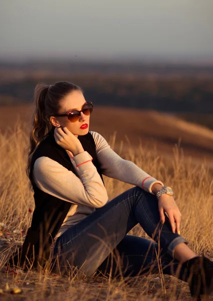 Bezauberndes, attraktives, seriöses Mädchen mit Sonnenbrille sitzt auf den Hügeln mit schöner Aussicht und beobachtet den Sonnenuntergang. — Stockfoto