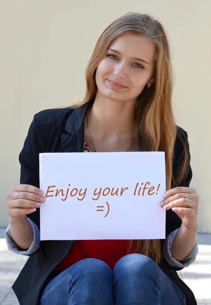 Beautiful blonde with the inscription on the white paper "Enjoy your life". Transcription.Record.Note.Enjoy your life.Beautiful, cute, nice, pretty blonde girl with long, straight hair . — стоковое фото