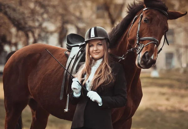 Professionele horsewoman. Professionele bedreven staan in de buurt van de bruine volbloed hengst. Blondie meisje met een speelse paard in zomer Horse Park. Paardrijden, staande in de buurt van de merrie in speciale uniform, vrouw met paard, gelukkig, stijl. Professioneel meisje Rider. — Stockfoto