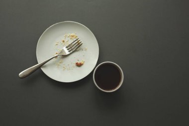 Empty plate with fork and crumbs and coffee cup after eating on gray background.