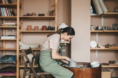 Kadın çömlekçi, çömlek çarkında vazo ve çömlek dolu raflara karşı kil stüdyoda çalışıyor..
