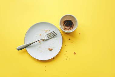 Empty plate with fork and crumbs after eating and cup with coffee grounds on yellow background.
