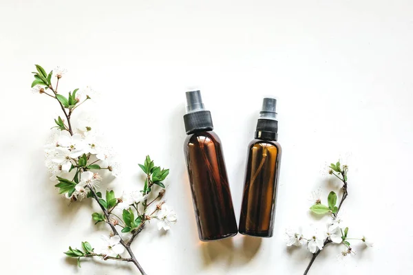 Butelki kosmetyczne wykonane z ciemnego bursztynowego szkła na białym tle z kwitnącymi gałęziami wiśni lub jabłoni — Zdjęcie stockowe