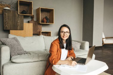 Gözlüklü, sıradan giyinmiş güzel bir kadın evde çalışırken dizüstü bilgisayar kullanıyor.. 