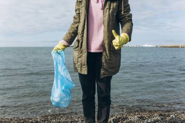 Kadın bir gönüllü, kirli bir sahilde çöp toplayan bir çöp torbasını elinde tutuyor.