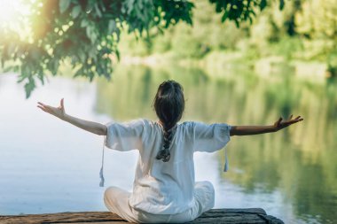 Göl kenarındaki doğada kendini keşfetme meditasyonu. Lotus pozisyonunda oturmuş, kollarını açmış meditasyon yapan bir kadın. 
