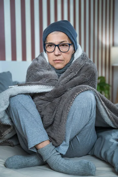 Senior woman with symptoms of seasonal flu, covered with blanket, sitting on the bed at home, feeling ill.