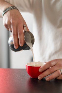 Kahveci kafede yeni demlenmiş kahve bardağına süt döküyor. Yumuşak köpük dokusunu, aromasını ve profesyonel sunumunu vurgular.