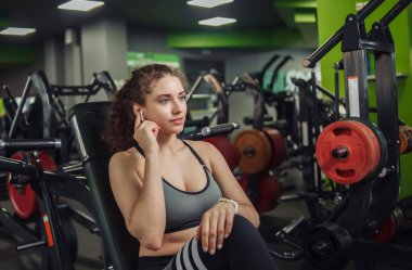 Spor salonundaki bankta oturan ve kulaklıklara dokunan genç ve fit bir kadın. Modern aletler. Egzersizler arasında dinlen..
