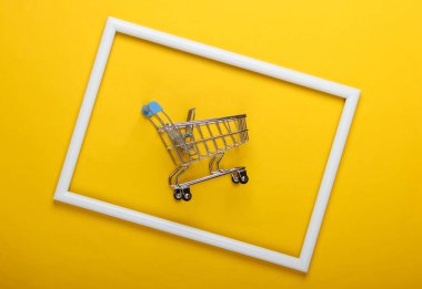 Beyaz çerçeveli sarı arka planda mini alışveriş arabası. Stüdyo çekimi. Yaratıcı düz yatak. Üst Manzara. Minimalizm