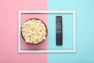 Beyaz çerçeveli pembe mavi pastel arka planda bir kase patlamış mısır ve televizyon kumandası. Boş zaman ve eğlence konsepti. Üst görünüm