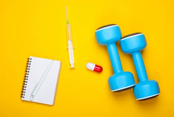 Pharmacology in sports. Dumbbell, capsule, syringe, notebook on yellow background. Vitamins, steroids. Top view