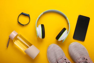 Spor ve spor aletlerinin düz kompozisyonu. Kablosuz kulaklık, akıllı bilezik, akıllı telefon, koşu ayakkabısı, sarı arka planda su şişesi. Üst görünüm