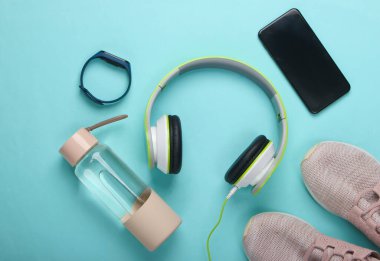 Spor ve spor aletlerinin düz kompozisyonu. Kablosuz kulaklık, akıllı bilezik, akıllı telefon, koşu ayakkabısı, mavi arka planda su şişesi. Üst görünüm