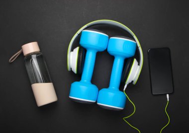 Düz yatak sporları, fitness kompozisyonu, aletler. Dambıllar, akıllı telefonlu kulaklıklar, siyah arka planda su şişesi. Üst görünüm