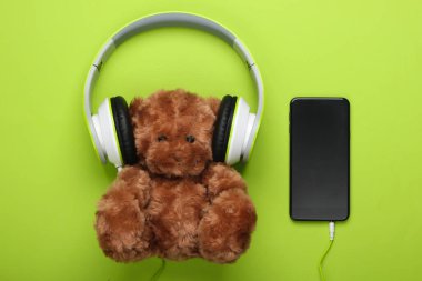 Müzik kulaklığı ve yeşil arka planda akıllı telefonu olan oyuncak ayı. Müzik aşığı konsepti, çocukluk. Üst görünüm