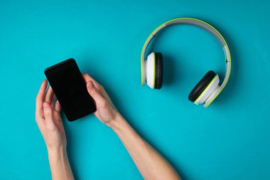 Kadın eller telefonu tutar ve kulaklıkları mavi arka planda senkronize eder. Modern aletler. Üst görünüm