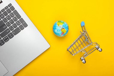 Küresel ağ, internet, online alışveriş, e-market. Laptop, küre ve sarı arka planda mini alışveriş arabası. Üst Manzara. Minimalizm