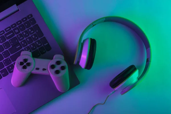 DJ or gaming concept. Laptop, headphones and gamepad with neon green purple light. Gradient glow. Top view