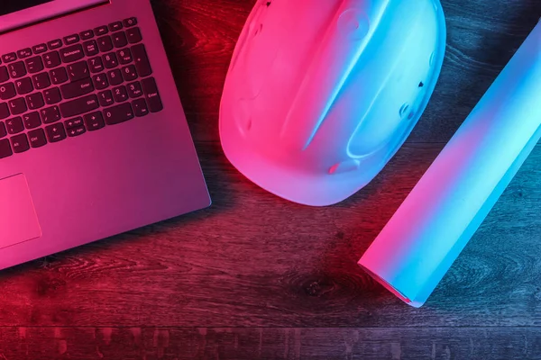 Engineering concept. Workspace. Construction helmet, roll of drawings, laptop on a wooden background with red-blue neon light. Top vew