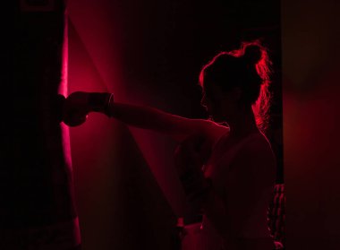 Siluetli, boks eldivenli kadın boksör. Koyu arkaplanda kırmızı ışıklı bir kum torbasına yumruk atıyor.