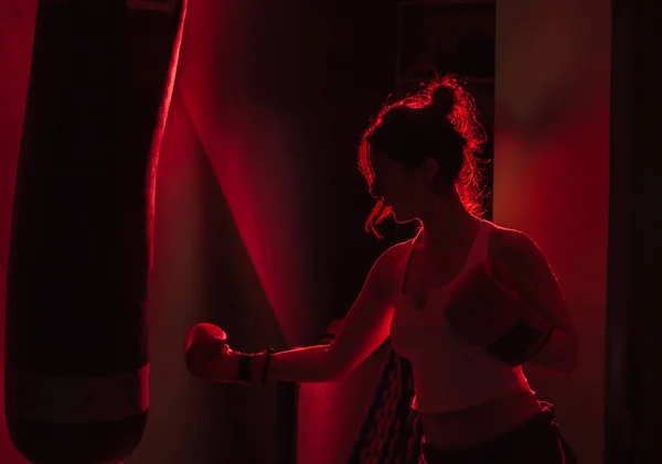 Silhouetted female boxer with boxing gloves punch a punching bag in red neon light over dark background