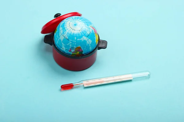 Global warming concept. Mini globe in a toy pan and thermometer on blue background. Minimalism. Climatic problems of our time.