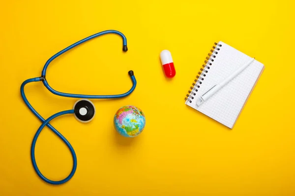 Global pandemic still life. Globe, stethoscope, capsule, notepad on a yellow background. Top view. Coronovirus outbreak