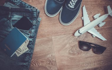 Seyahat ya da turizm konsepti. Kot pantolon, yüz dolarlık banknotlar, spor ayakkabılar, akıllı telefon, pasaport, güneş gözlüğü, ahşap zeminde bir uçak. Üst Manzara. Düz yatıyordu