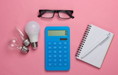 Enerji tasarrufu. Ampul, gözlük, pembe mavi pastel arka planda not defteri olan hesap makinesi. Üst görünüm