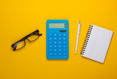 Hesap makinesi ve gözlük, sarı arka planda defter. Sekreter, ekonomist ya da ofis çalışanı konsepti. Üst görünüm