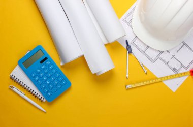 İnşaat kaskı, plan ruloları, proje planı ve sarı arka planda hesap makinesi. Bir ev inşa etmenin maliyetinin hesaplanması. Üst Manzara. Düz yatıyordu