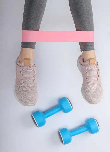 Female legs in sports leggings and sneakers doing exercises with fitness elastic on white background with dumbbells. Top view