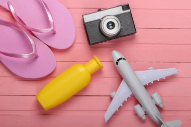 Fotoğraf makinesi, uçak, pembe ahşap arka planda güneş kremi olan parmak arası terlikler. Plaj aksesuarları